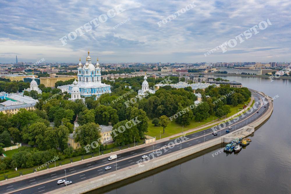 На фотографии изображён Смольный собор, один из шедевров архитектуры елизаветинского барокко, расположенный в окружении парка. На переднем плане видна набережная Невы и современная дорога, гармонично вписывающаяся в городской пейзаж. Снимок передаёт спокойствие и величие города в пасмурную погоду.